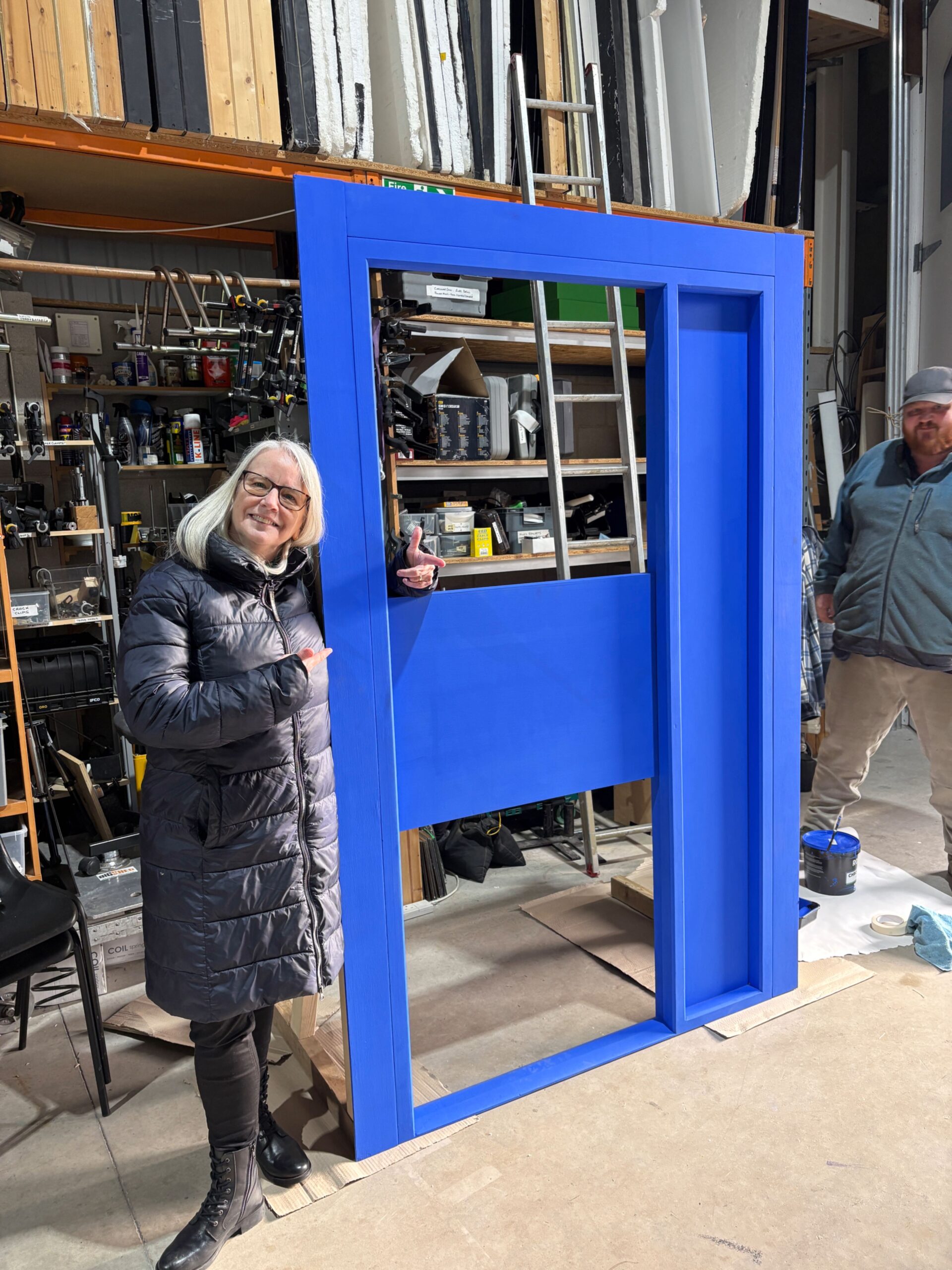 Smiling woman in winter coat giving thumbs up beside large blue wooden structure in a workshop.