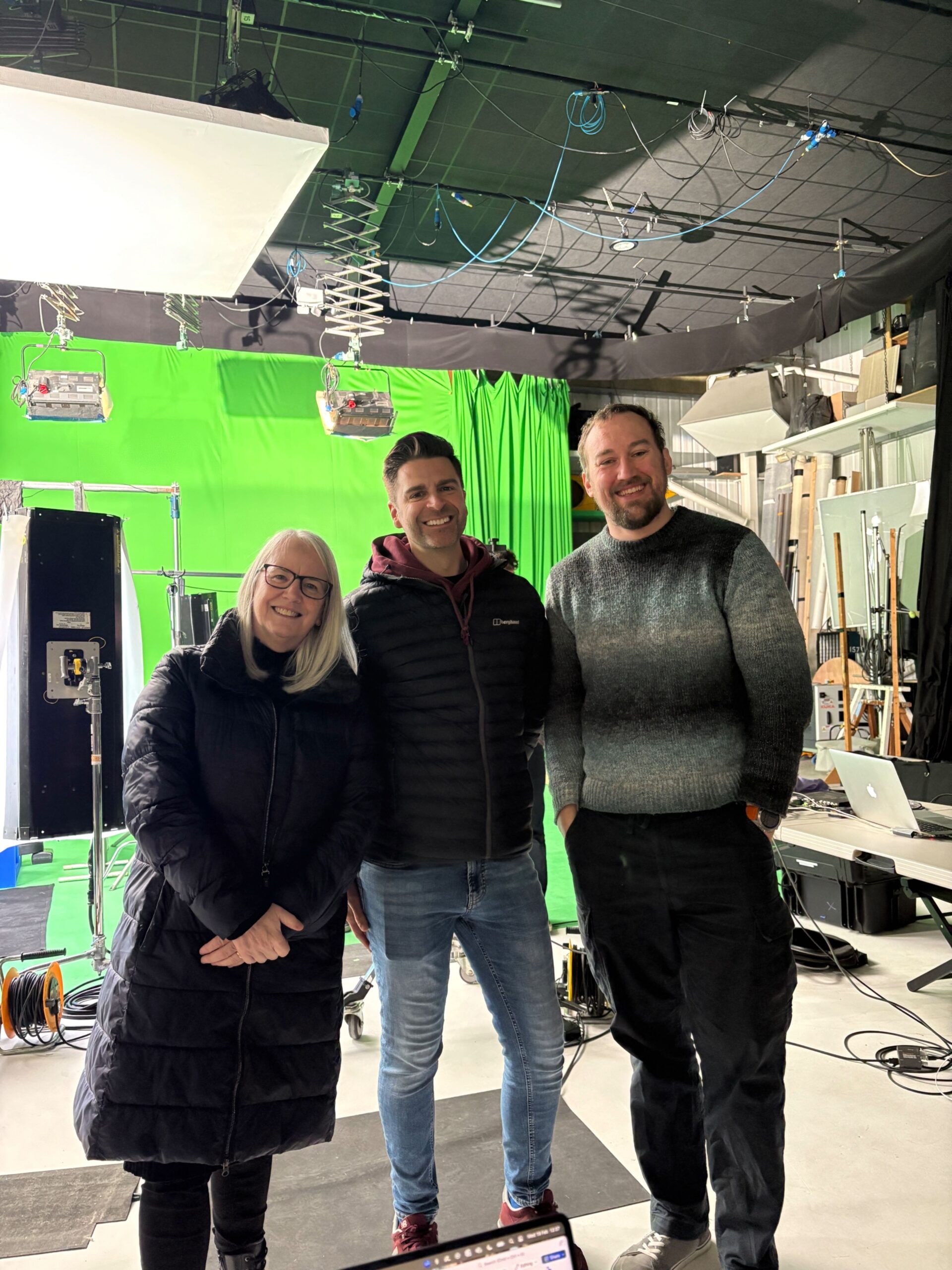 Three people smiling in a studio with green screen and lighting equipment in the background.