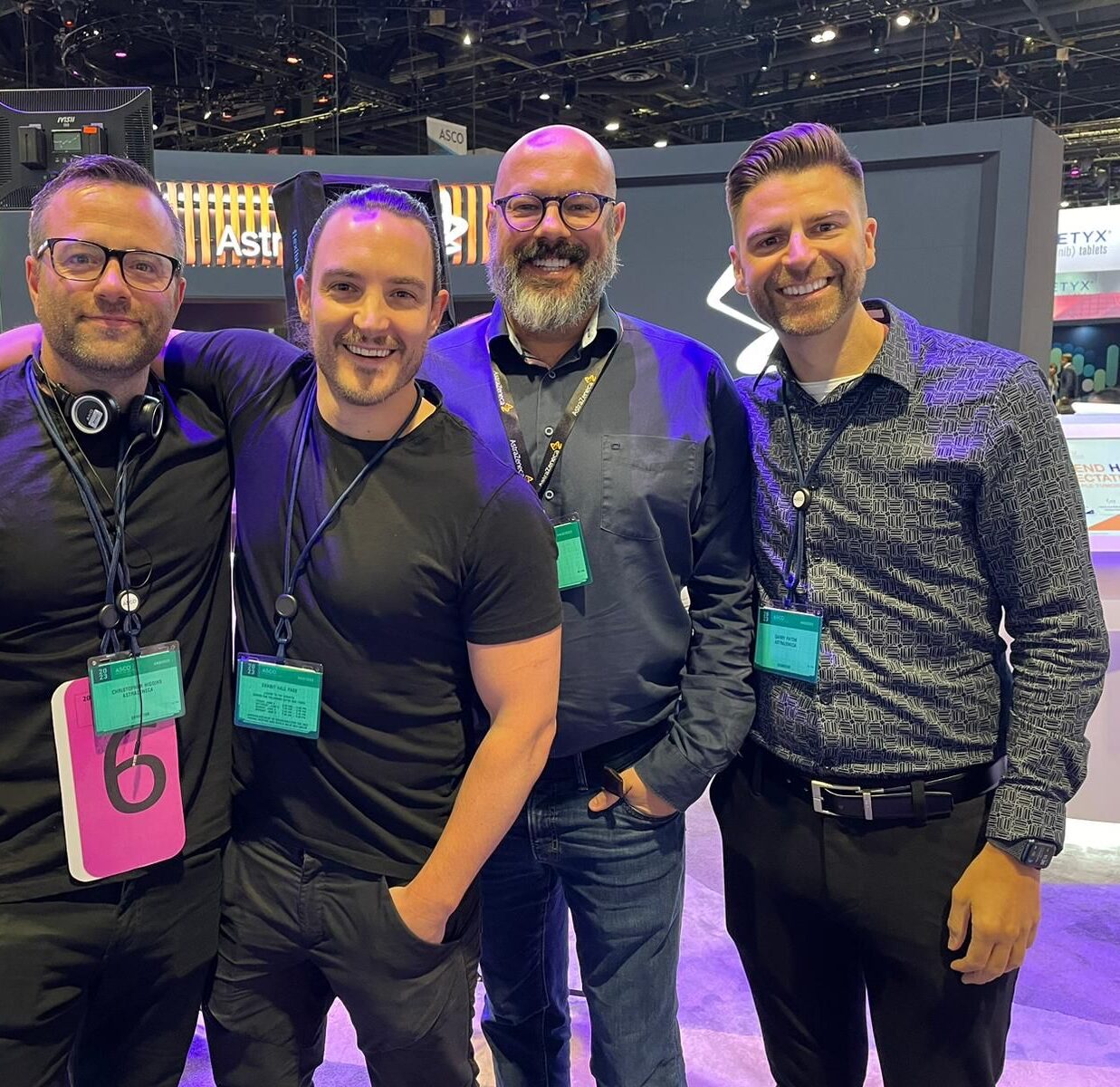 Four men smiling and standing together on a convention floor, wearing exhibitor badges at ASCO 2023 with cameras and branded booths in the background.