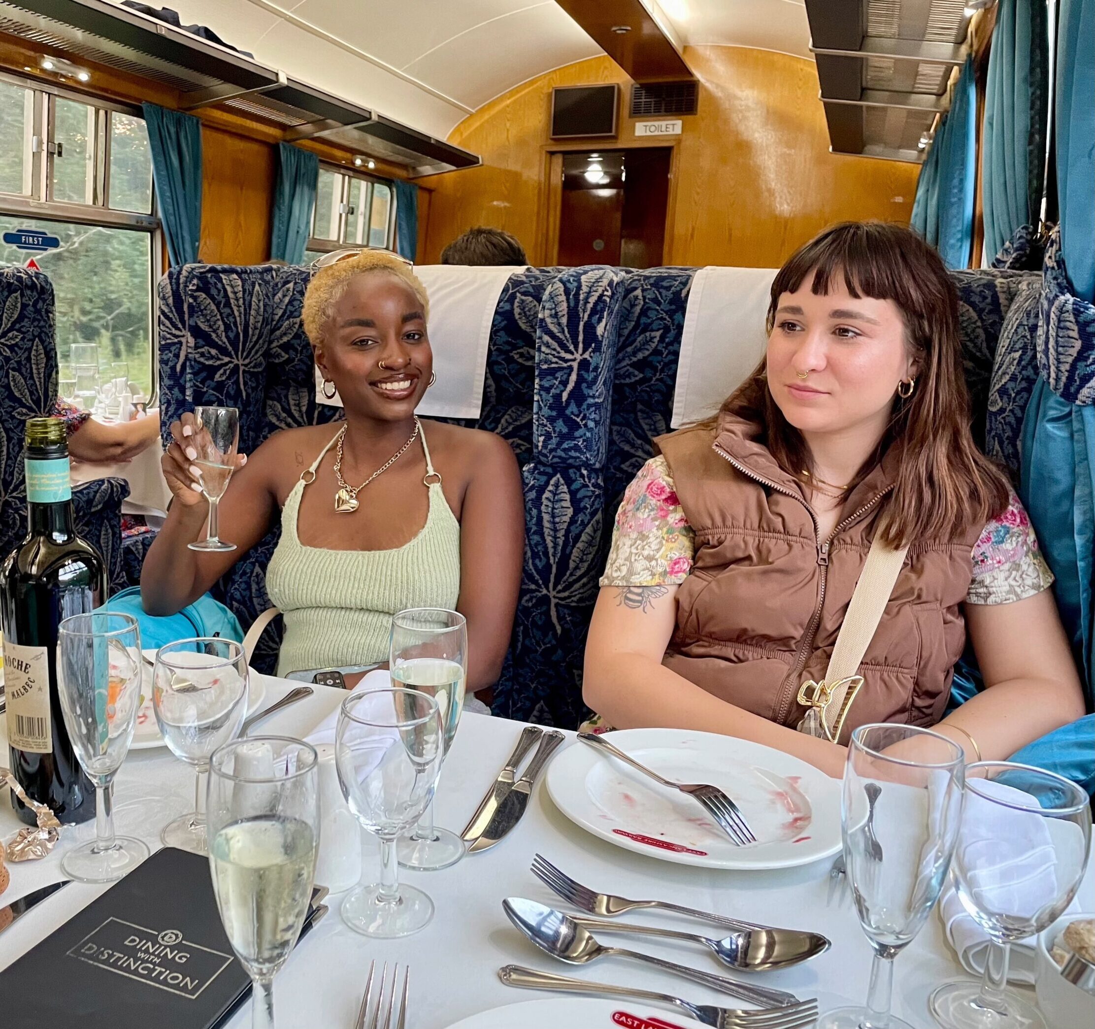 Two women seated at a dining table on a vintage train, surrounded by glassware and bottles, enjoying a meal in a formal dining carriage.