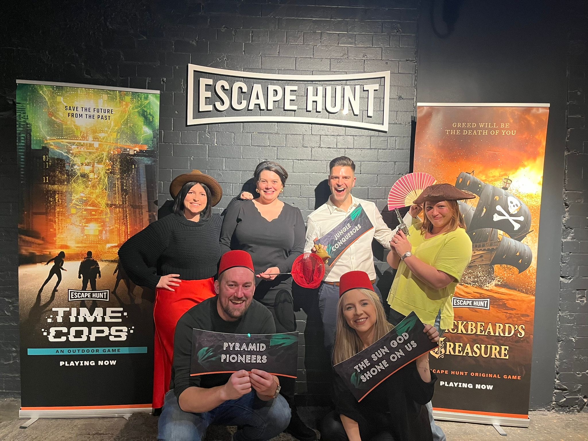 Team posing after an escape room challenge, holding props and signs in front of themed posters for Time Cops and Blackbeard’s Treasure.